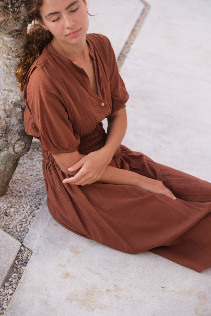 Presenting our Brown Handwoven Midi Dress, showcased by a model next to a tree. This dress highlights detailed handcrafting and puff sleeves, adding a sophisticated touch to your daily wardrobe. Made from soft, breathable cotton with sustainable practices, it seamlessly blends style and comfort.