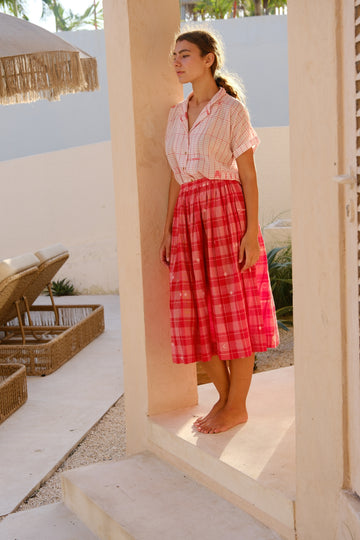 Presenting the Crimson Checkered Cuban Shirt, a joint creation by Audrey E Leary and World of Crow. This shirt, crafted from delicately hand-woven jamdani fabric, features a soft pink hue adorned with red patterns. With its short sleeves and airy, lightweight design, it’s ideal for daily wear. Sustainably produced from plush cotton, it pairs beautifully with a red checkered skirt