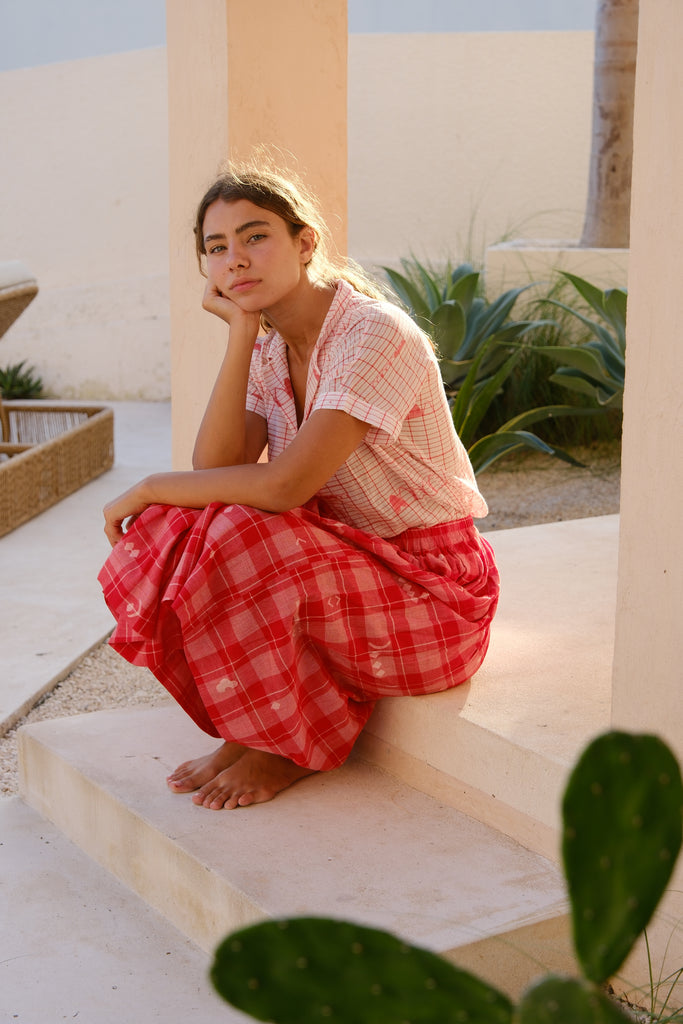 Presenting the Crimson Checkered Cuban Shirt, a joint creation by Audrey E Leary and World of Crow. This shirt, crafted from delicately hand-woven jamdani fabric, features a soft pink hue adorned with red patterns. With its short sleeves and airy, lightweight design, it’s ideal for daily wear. Sustainably produced from plush cotton, it pairs beautifully with a red checkered skirt