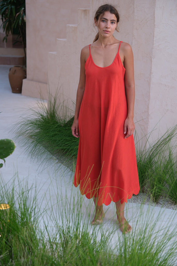 Presenting the Holiday Slip Dress, a collaboration between Audrey E Leary and World of Crow. This sleeveless, orangish-red midi dress with scalloped details, crafted from handwoven cotton, is lightweight and breathable, making it a versatile and sustainable choice for layering and outings.