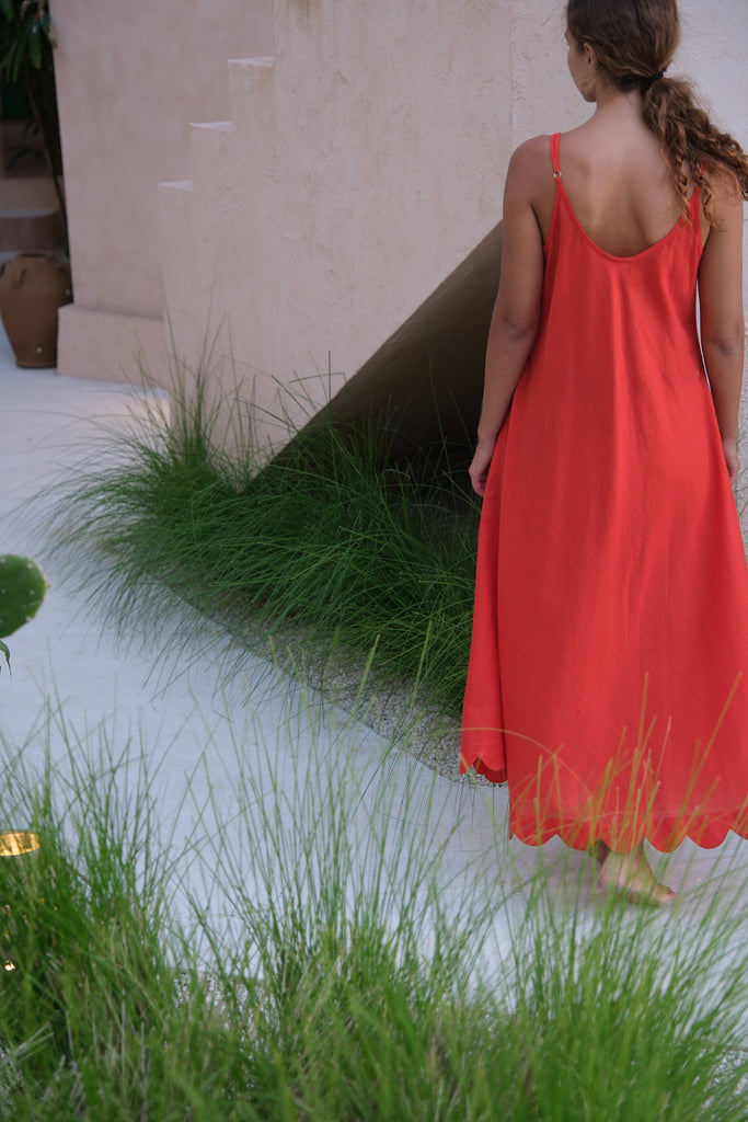 Presenting the Holiday Slip Dress, a collaboration between Audrey E Leary and World of Crow. This sleeveless, orangish-red midi dress with scalloped details, crafted from handwoven cotton, is lightweight and breathable, making it a versatile and sustainable choice for layering and outings.