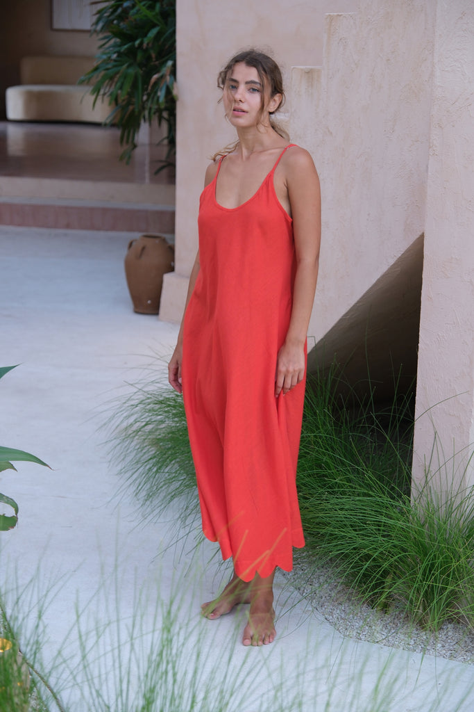 Presenting the Holiday Slip Dress, a collaboration between Audrey E Leary and World of Crow. This sleeveless, orangish-red midi dress with scalloped details, crafted from handwoven cotton, is lightweight and breathable, making it a versatile and sustainable choice for layering and outings.