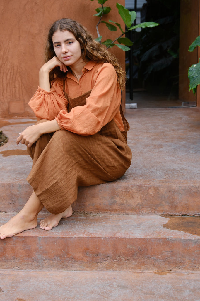 Presenting the Work-Boy’s Dungarees, crafted from handwoven cotton with retro self-check patterns and elegant pockets from the Audrey X Crow Collection. Shown on a woman in an orange shirt and brown dungarees sitting on steps, these dungarees offer a blend of classic craftsmanship and vintage charm, merging comfort with style.