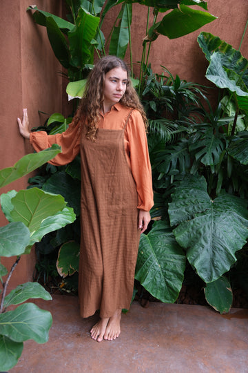 Presenting the Work-Boy’s Dungarees, crafted from handwoven cotton with retro self-check patterns and elegant pockets from the Audrey X Crow Collection. Shown on a woman in an orange shirt and brown dungarees sitting on steps, these dungarees offer a blend of classic craftsmanship and vintage charm, merging comfort with style.